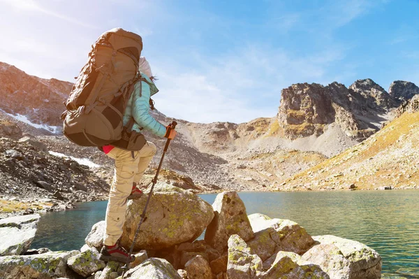Una ragazza escursionista in occhiali da sole con uno zaino e bastoni di monitoraggio sale a una roccia alta sullo sfondo di rocce e un lago di alta montagna — Foto Stock