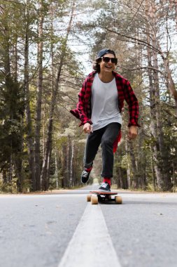 Bir kap ve ekose gömlekli bir genç gülmek hipster onun longboard ormanın içinde bir ülke yolda sürme