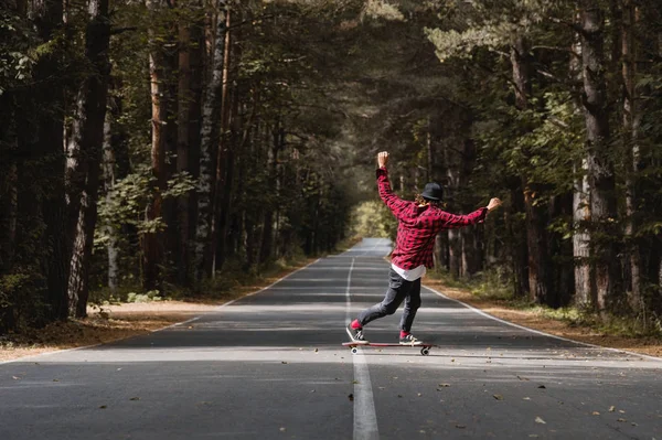 Um jovem hipster com um boné e camisa xadrez está montando seu longboard em uma estrada rural na floresta — Fotografia de Stock