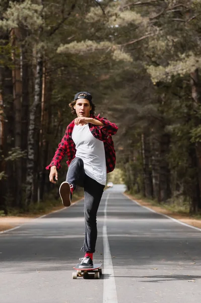 Ein junger Hipster mit Schirmmütze und kariertem Hemd fährt mit seinem Longboard auf einer Landstraße im Wald — Stockfoto