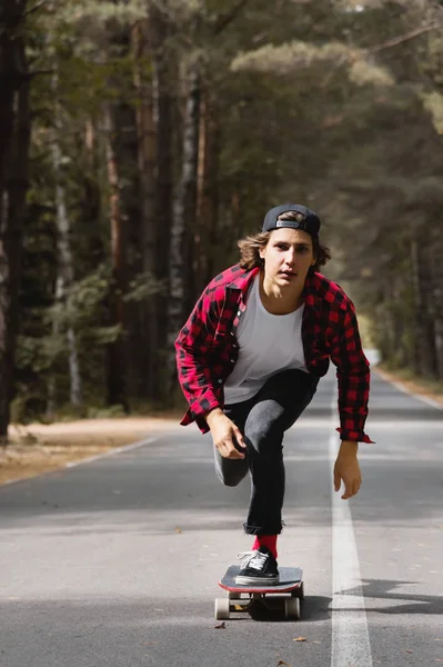 모자와 격자 무늬 셔츠에 젊은 hipster 숲에 국가로에 그의 longboard 타고 — 스톡 사진