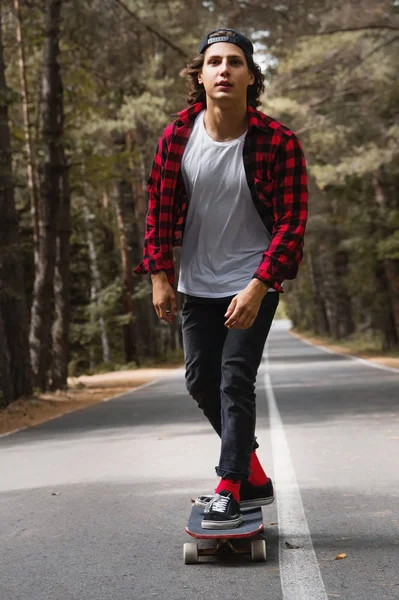 Um jovem hipster com um boné e camisa xadrez está montando seu longboard em uma estrada rural na floresta — Fotografia de Stock