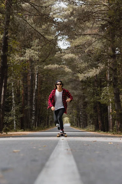 Un joven hipster con gorra y camisa a cuadros está montando su longboard en un camino rural en el bosque — Foto de Stock