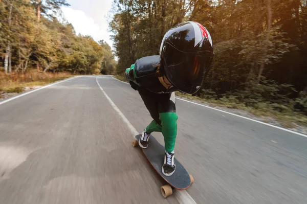 Um jovem cavaleiro em um terno de couro especial e um passeio de meio capacete em alta velocidade em seu longboard em uma estrada rural na floresta — Fotografia de Stock