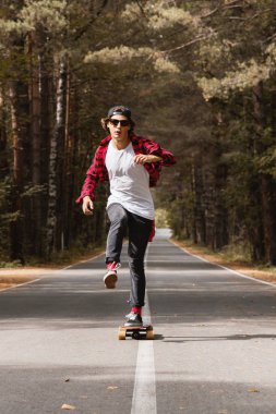 Bir kap ve ekose gömlek genç bir hipster onun longboard ormanın içinde bir ülke yolda sürme
