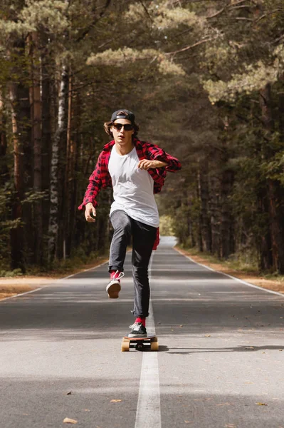 Un joven hipster con gorra y camisa a cuadros está montando su longboard en un camino rural en el bosque — Foto de Stock