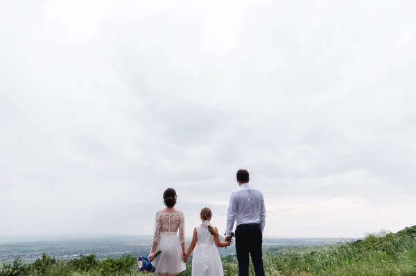 Giovane coppia sposata e la loro piccola figlia il giorno del matrimonio sono in piedi nella natura tenendosi per mano — Foto Stock