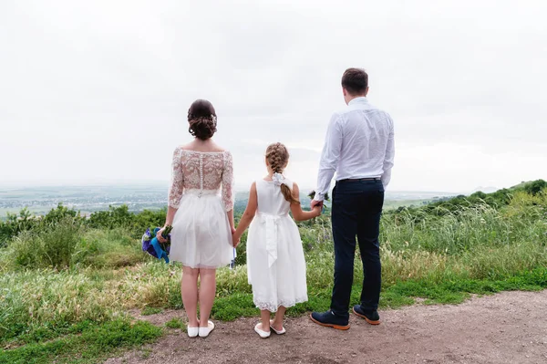 Joven pareja casada y su hija pequeña en el día de la boda están de pie en la naturaleza de la mano — Foto de Stock