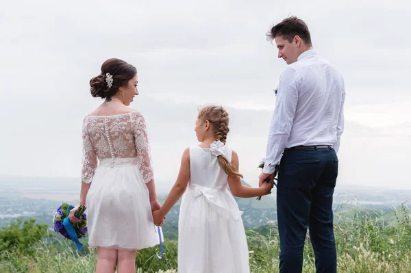 Jeune couple marié et leur petite fille le jour du mariage sont debout dans la nature tenant la main — Photo