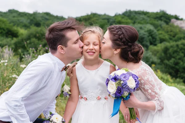 Jovens pais em vestidos de noiva beijam sua jovem filha em bochechas — Fotografia de Stock