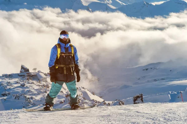 Um snowboarder profissional com uma mochila está na neve no alto das montanhas contra o fundo de nuvens baixas e as montanhas do Cáucaso durante o pôr do sol — Fotografia de Stock
