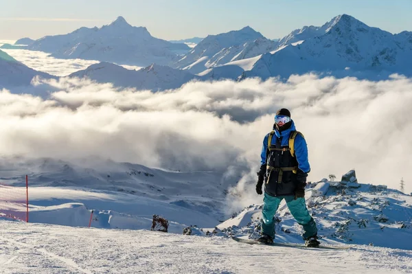 Een professionele snowboarder met een rugzak staat op de hoge sneeuw in de bergen tegen de achtergrond van de lage wolken en de bergen van de Kaukasus tijdens de ondergaande zon — Stockfoto