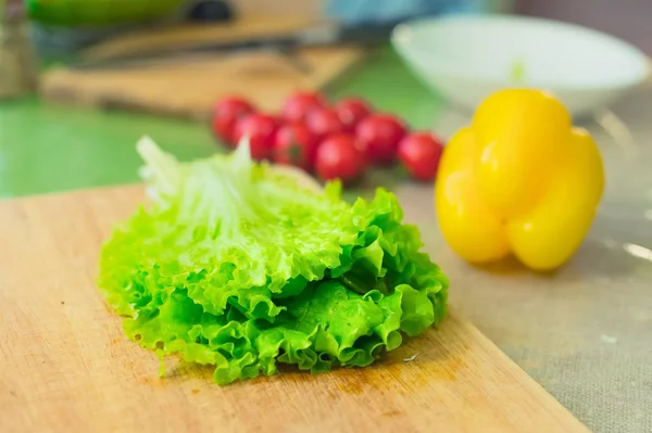 Hojas de lechuga verde fresco se encuentran en una tabla de cortar de madera junto al pimiento amarillo y tomates rojos cereza en una mesa verde — Foto de Stock