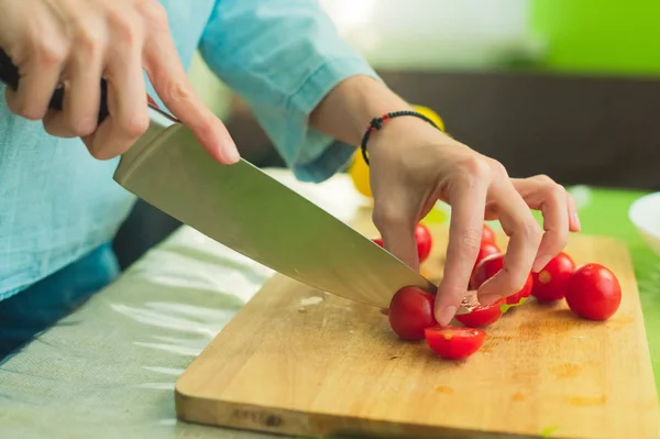 Руки молодой девушки рубят помидоры черри на деревянной разделочной доске на зеленом столе в домашних условиях — стоковое фото