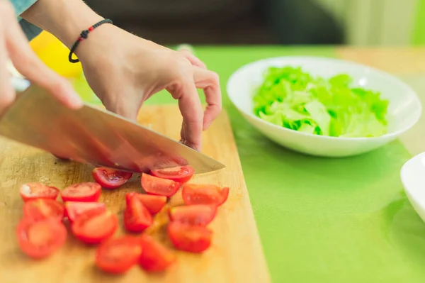 Hands of a young girl chop the cherry tomatoes on a wooden cutting board on a green table in a home setting — Stock Photo, Image