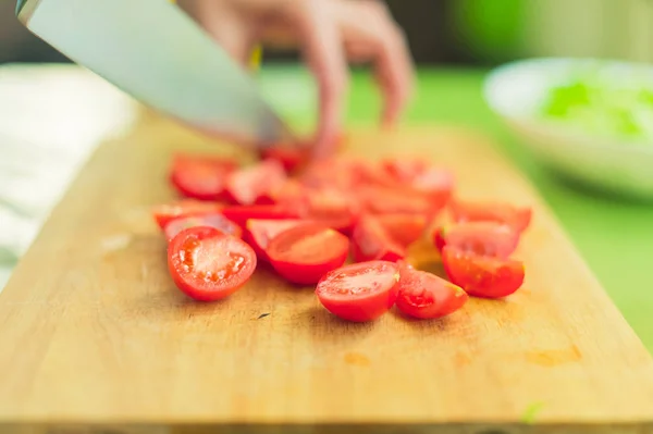 Las manos de una joven cortan los tomates cherry en una tabla de cortar de madera sobre una mesa verde en un entorno hogareño — Foto de Stock