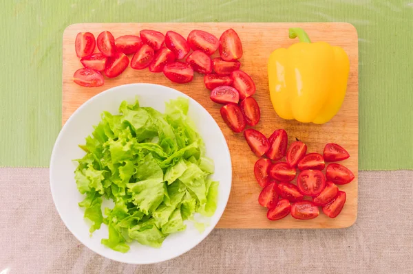 Hojas frescas de lechuga verde picada se encuentran en una tabla de cortar de madera junto al pimiento amarillo y tomates rojos picados de cereza en una mesa verde — Foto de Stock
