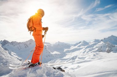 Bir kayakçı bir turuncu bir sırt çantası onun ile genel geri kask giymiş ve elinde Kayak Polonyalılar ile güzel bir arka planda karlı bir uçurum önünde bir uçurumun üzerinde duruyor