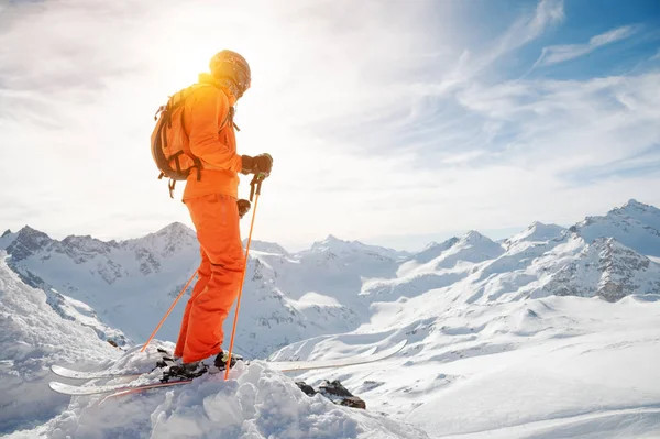 Lyžař v pomeranč celkově s batohem na jeho zpět nosit helmu a s lyžařské hole v ruce stojí na svahu před zasněžené propasti na pozadí krásné — Stock fotografie