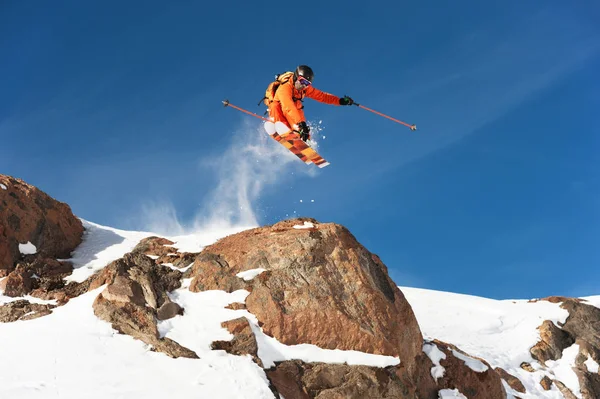 En professionell skidåkare gör en jump-släpp från en hög klippa mot en blå himmel som lämnar ett spår av snow pulver i bergen — Stockfoto