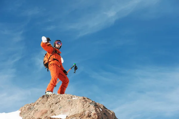Porträt eines Skifahrers in orangefarbenem Overall mit Rucksack auf dem Rücken und Skiern auf den Schultern in Helm steht auf einem Felsen vor blauem Himmel. — Stockfoto