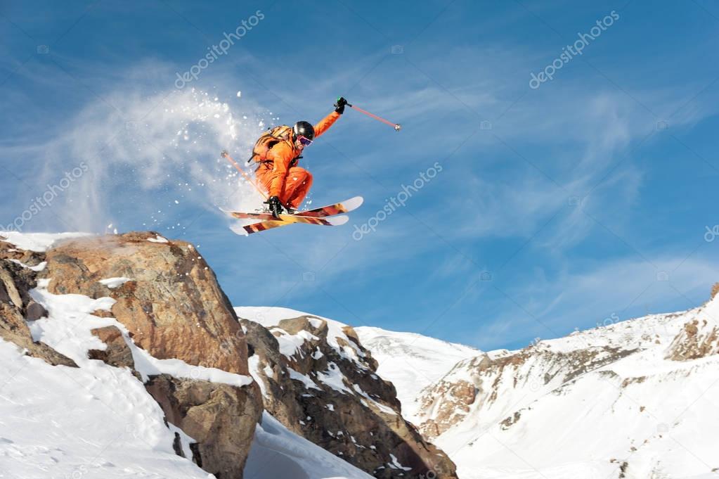 A professional skier makes a jump-drop from a high cliff against a blue sky leaving a trail of snow powder in the mountains