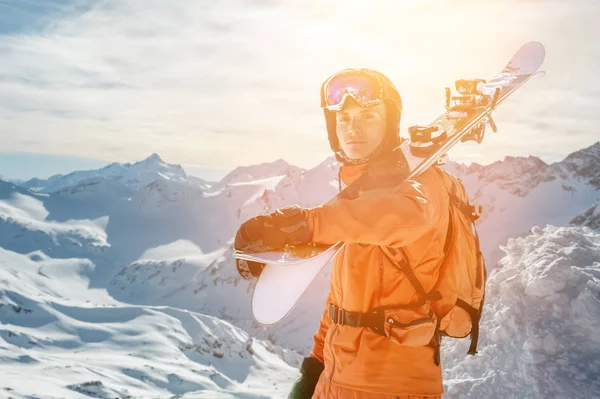 Portrait of a Skier in an orange overall with a backpack on his back and skis on his shoulders in a helmet stands on a rock against the blue sky. — Stock Photo, Image