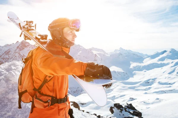 Retrato de um esquiador em um geral laranja com uma mochila em suas costas e esquis em seus ombros em um capacete fica em uma rocha contra o céu azul . — Fotografia de Stock