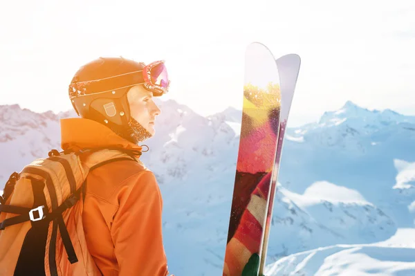 A skier in an orange overall with a backpack on his back wearing a helmet and with ski poles in his hands is standing on a precipice in front of a snowy abyss in the background of a beautiful — Stock Photo, Image