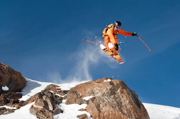 En professionell skidåkare gör en jump-släpp från en hög klippa mot en blå himmel som lämnar ett spår av snow pulver i bergen — Stockfoto