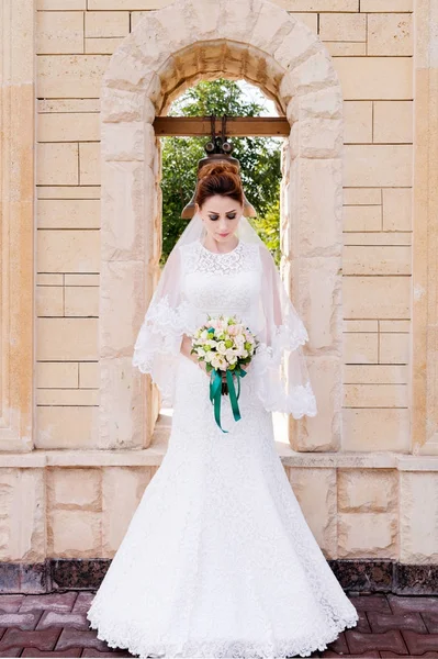 Красива наречена в весільній сукні і весільний букет в руках стоїть на відкритому повітрі на фоні кам'яної арки з дзвінком. Концепція весілля щастя — стокове фото