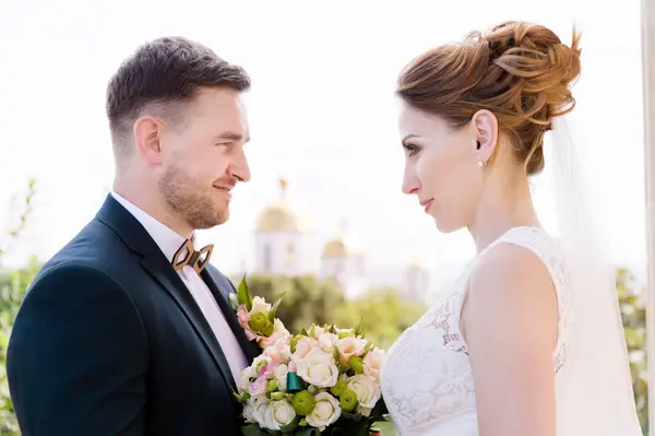 Um belo casal de recém-casados em um dia de casamento com um buquê na mão contra o fundo de uma igreja cristã ortodoxa . — Fotografia de Stock