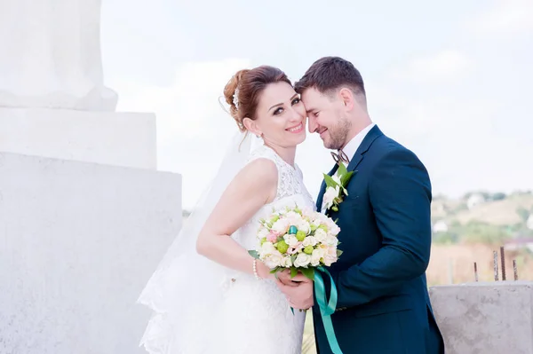 Retrato de um lindo casal lua de mel em um dia de casamento com um buquê na mão contra o fundo de um monumento cristão ortodoxo com anjos . — Fotografia de Stock