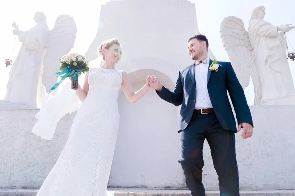 Retrato de um lindo casal lua de mel em um dia de casamento com um buquê na mão contra o fundo de um monumento cristão ortodoxo com anjos . — Fotografia de Stock