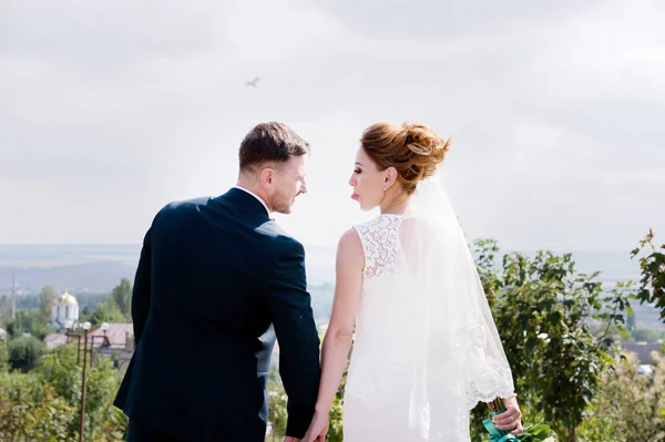 Portrait d'un beau couple en lune de miel le jour d'un mariage avec un bouquet dans les mains se regardant rire et sourire contre une allée verte à l'extérieur . — Photo
