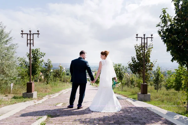 Retrato de um casal encantador lua de mel em um dia de casamento com um buquê em mãos olhando umas para as outras rindo e sorrindo contra um beco verde ao ar livre . — Fotografia de Stock