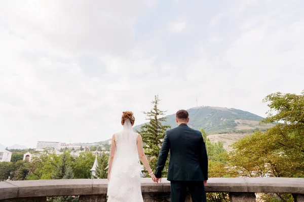 Milující pár novomanželů stojan drží za ruce a všeobjímající na pozadí krajiny města resort město v severním Kavkazu. — Stock fotografie