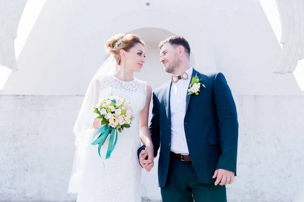 Retrato de um lindo casal lua de mel em um dia de casamento com um buquê na mão contra o fundo de um monumento cristão ortodoxo com anjos . — Fotografia de Stock