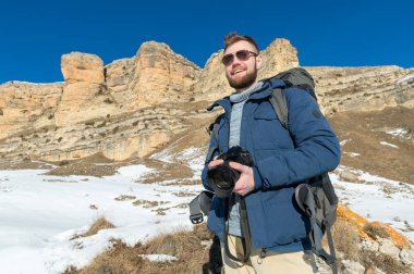 Portre A sakallı hippi fotoğrafçı bir sırt çantası ve onun omuz standları epik kayalar arka planı elinde bir Dslr fotoğraf makinesi ile üzerinde büyük bir sırt çantası ile giyen güneş gözlüğü