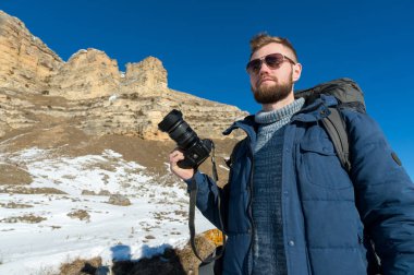 Portre A sakallı hippi fotoğrafçı bir sırt çantası ve onun omuz standları epik kayalar arka planı elinde bir Dslr fotoğraf makinesi ile üzerinde büyük bir sırt çantası ile giyen güneş gözlüğü