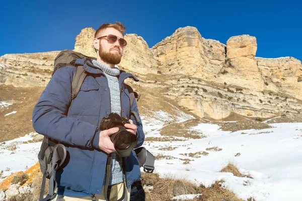 Ritratto Un fotografo hipster barbuto con uno zaino e occhiali da sole con un grande zaino sulle spalle si erge con una fotocamera DSLR tra le mani sullo sfondo di rocce epiche — Foto Stock