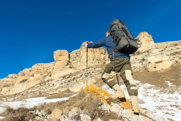 Batohem s velkým batohem a hole stoupá ke skále na pozadí epické skal v zimní sezóně. — Stock fotografie
