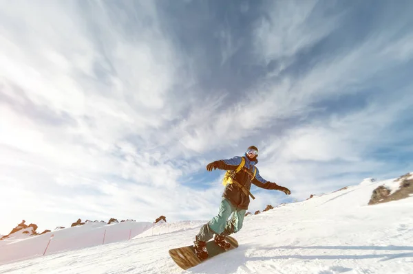 Ένας snowboarder σε μια μάσκα σκι και ένα σακίδιο είναι ιππασία σε μια πλαγιά χιονισμένη αφήνοντας πίσω μια σκόνη χιόνι κατά το γαλάζιο του ουρανού και τη ρύθμιση Κυρ — Φωτογραφία Αρχείου