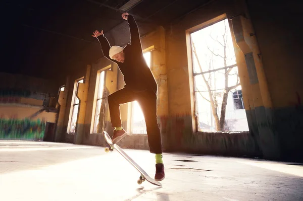 A young skater in a white hat and a black sweatshirt does a trick with a skate jump in an abandoned building in the backlight of the setting sun.
