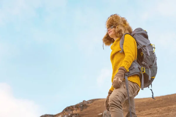 La mochilera con gafas de sol y un gran sombrero de piel del norte con una mochila en la espalda está de pie sobre una roca y mirando hacia los acantilados que se esconden en la roca . — Foto de Stock