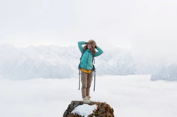 Een gelukkig lachend hipster meisje in een donsjack met een rugzak reist en in een grote vacht hoed staat op de top van een rots — Stockfoto
