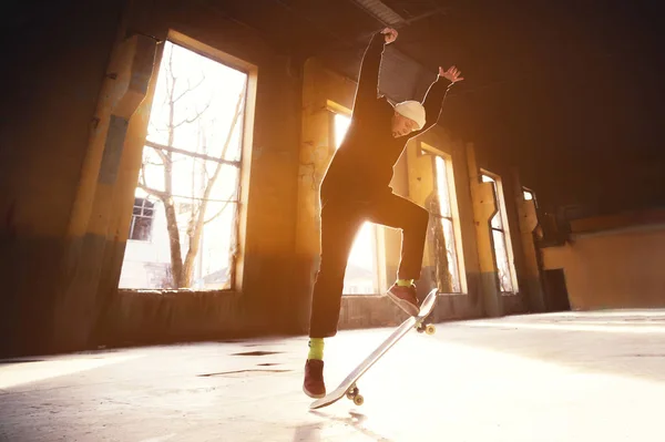 Een jonge schaatser in een witte hoed en een zwarte trui doet een truc met een skate-sprong in een verlaten gebouw in de achtergrondverlichting van de ondergaande zon. — Stockfoto