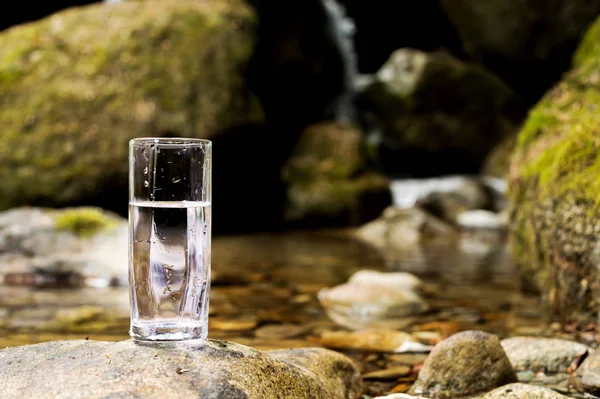 Un vetro trasparente con acqua minerale del fiume di montagna sorge su una pietra accanto al torrente del fiume di montagna — Foto Stock