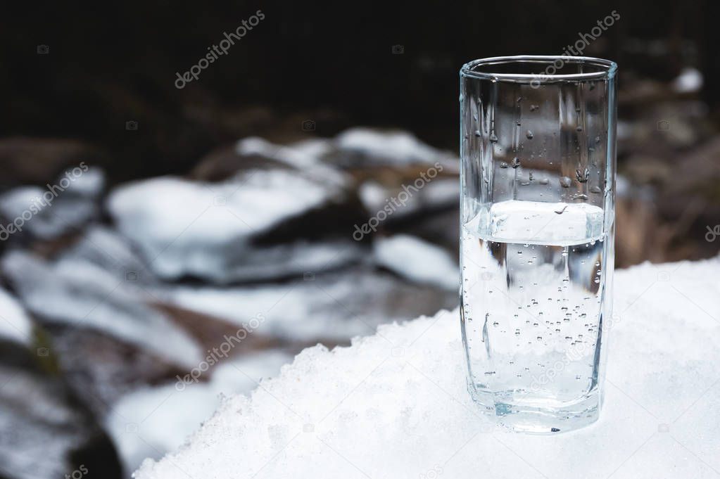 A transparent glass glass with drinking mountain water stands in the snow against a background of a clean mountain river and a forest in winter.