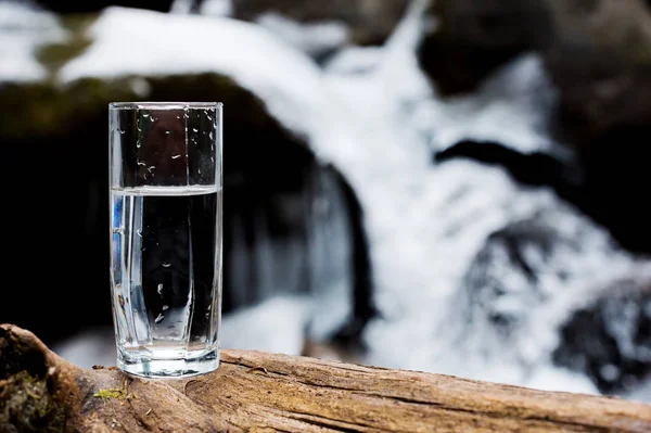 Un vetro trasparente con acqua minerale di montagna potabile sorge sul legno sullo sfondo di un flusso rapido di un fiume di montagna pulito in inverno. . — Foto Stock